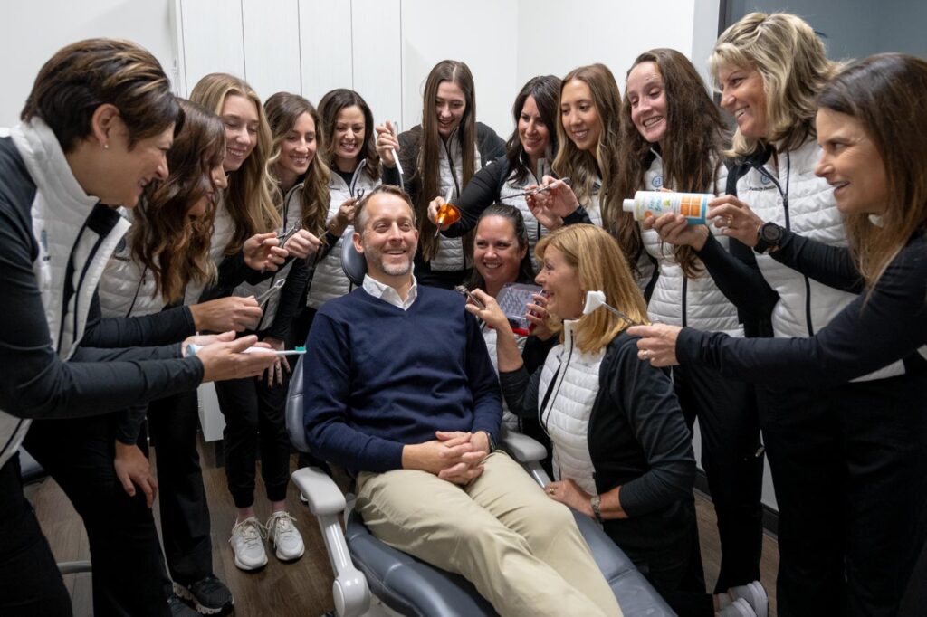 A man sits, smiling, in a dentist chair while a group of women surround him, playfully holding dental tools, hair products, and grooming items toward his head. Everyone is laughing and having funâ€”Contact Ryan R Love DDS to join the fun!.